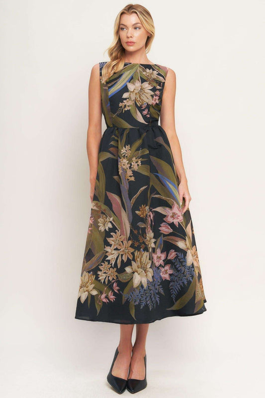 A woman modeling a sleeveless midi dress with a dark navy base and a detailed botanical print in beige, green, and pink, paired with black pointed-toe heels against a plain white studio background.