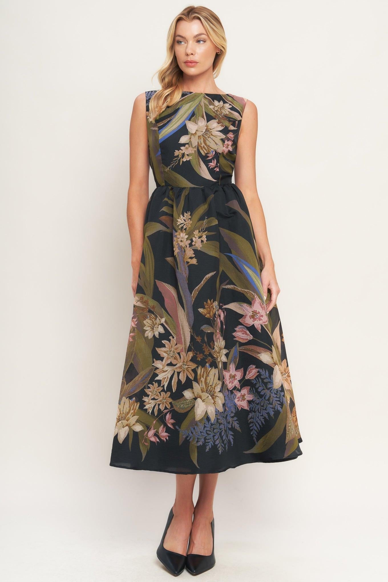 A woman modeling a sleeveless midi dress with a dark navy base and a detailed botanical print in beige, green, and pink, paired with black pointed-toe heels against a plain white studio background.