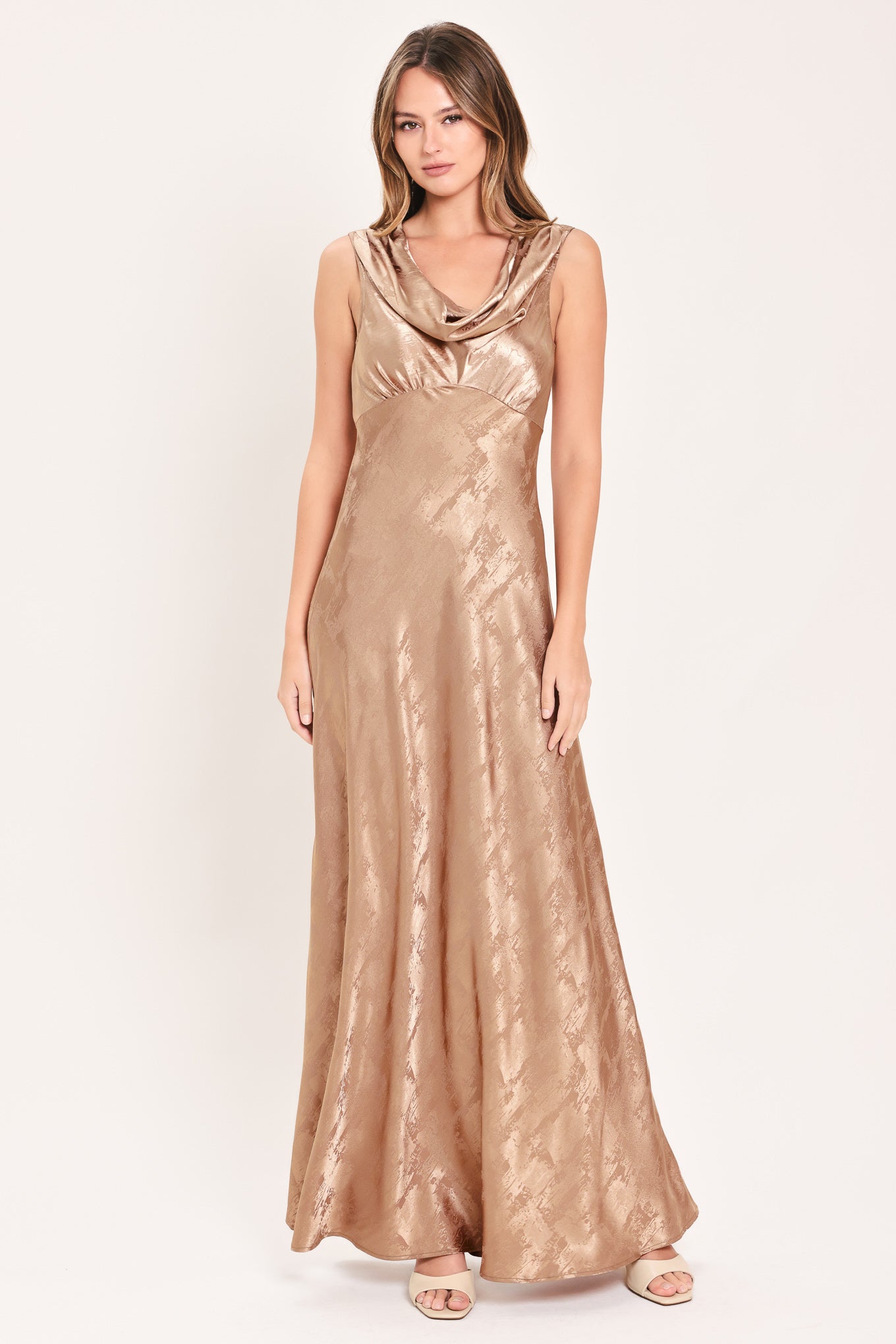 A woman modeling a sleeveless, floor-length metallic champagne-gold gown with a draped cowl neckline and a subtle tonal pattern, set against a plain light background.