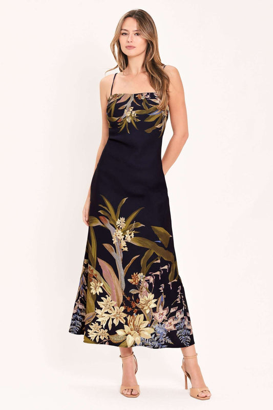 A woman modeling a sleeveless navy maxi dress with a tropical floral and botanical print in beige, gold, and muted green, paired with nude high-heeled sandals against a plain white studio background.