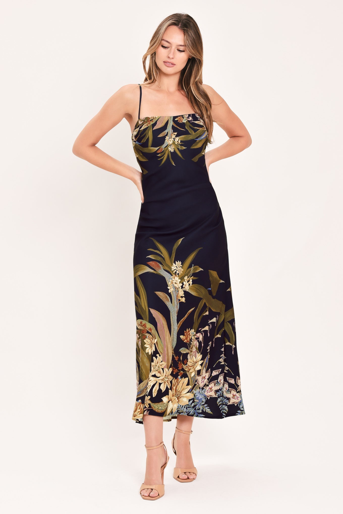 A woman modeling a sleeveless midi dress with thin straps, featuring a dark navy base with a tropical floral and botanical print in olive green, beige, and light blue, set against a plain off-white studio background.