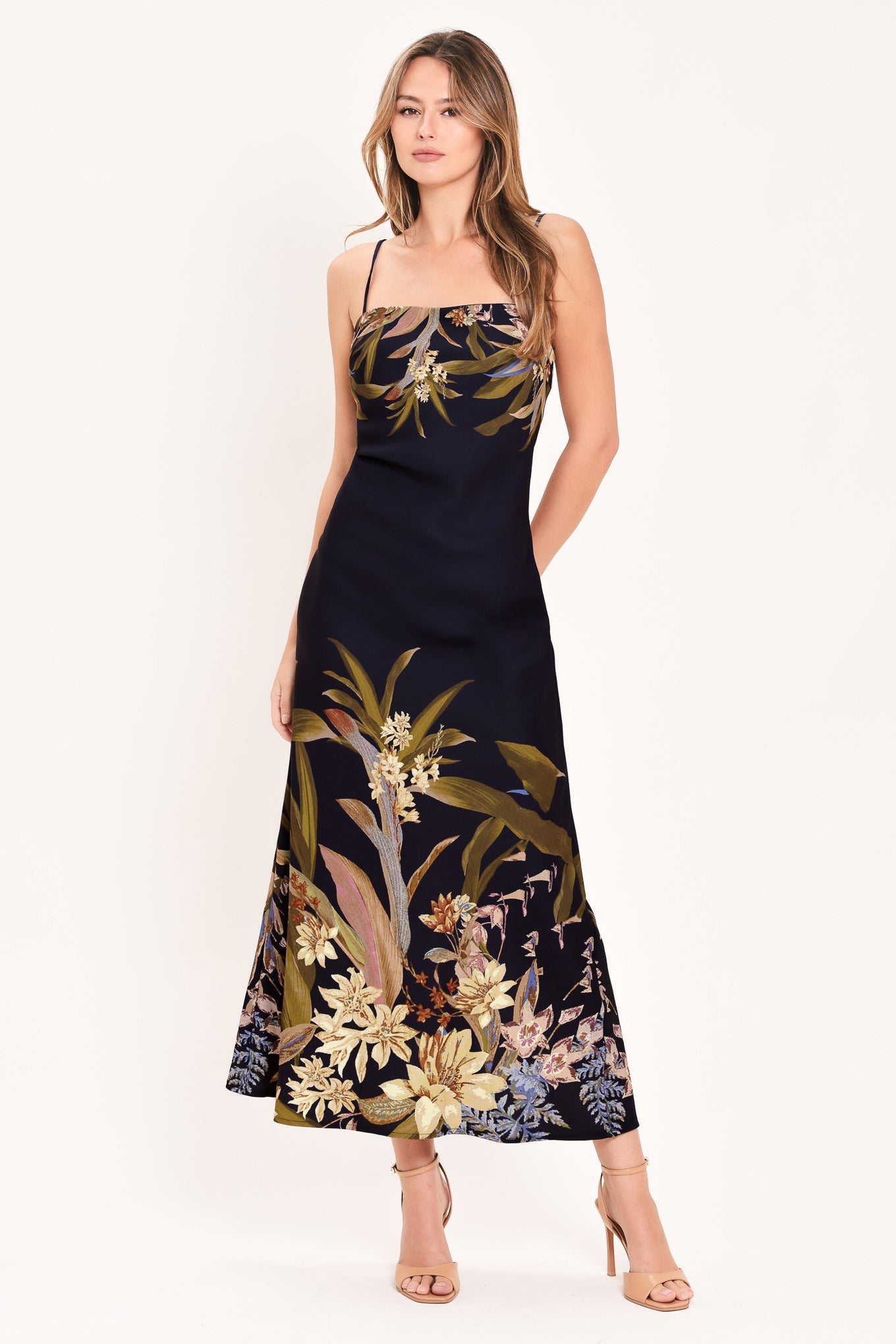 A woman modeling a sleeveless navy maxi dress with a tropical floral and botanical print in beige, gold, and muted green, paired with nude high-heeled sandals against a plain white studio background.