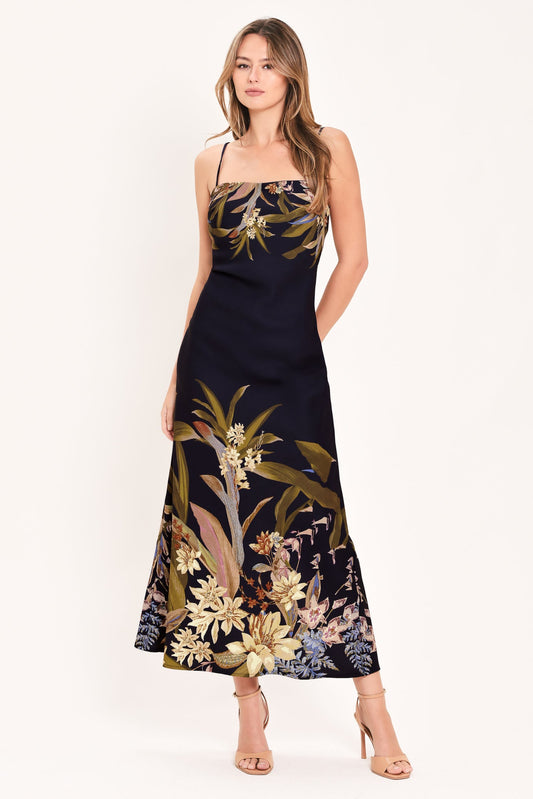A woman modeling a sleeveless navy maxi dress with a tropical floral and botanical print in beige, gold, and muted green, paired with nude high-heeled sandals against a plain white studio background.