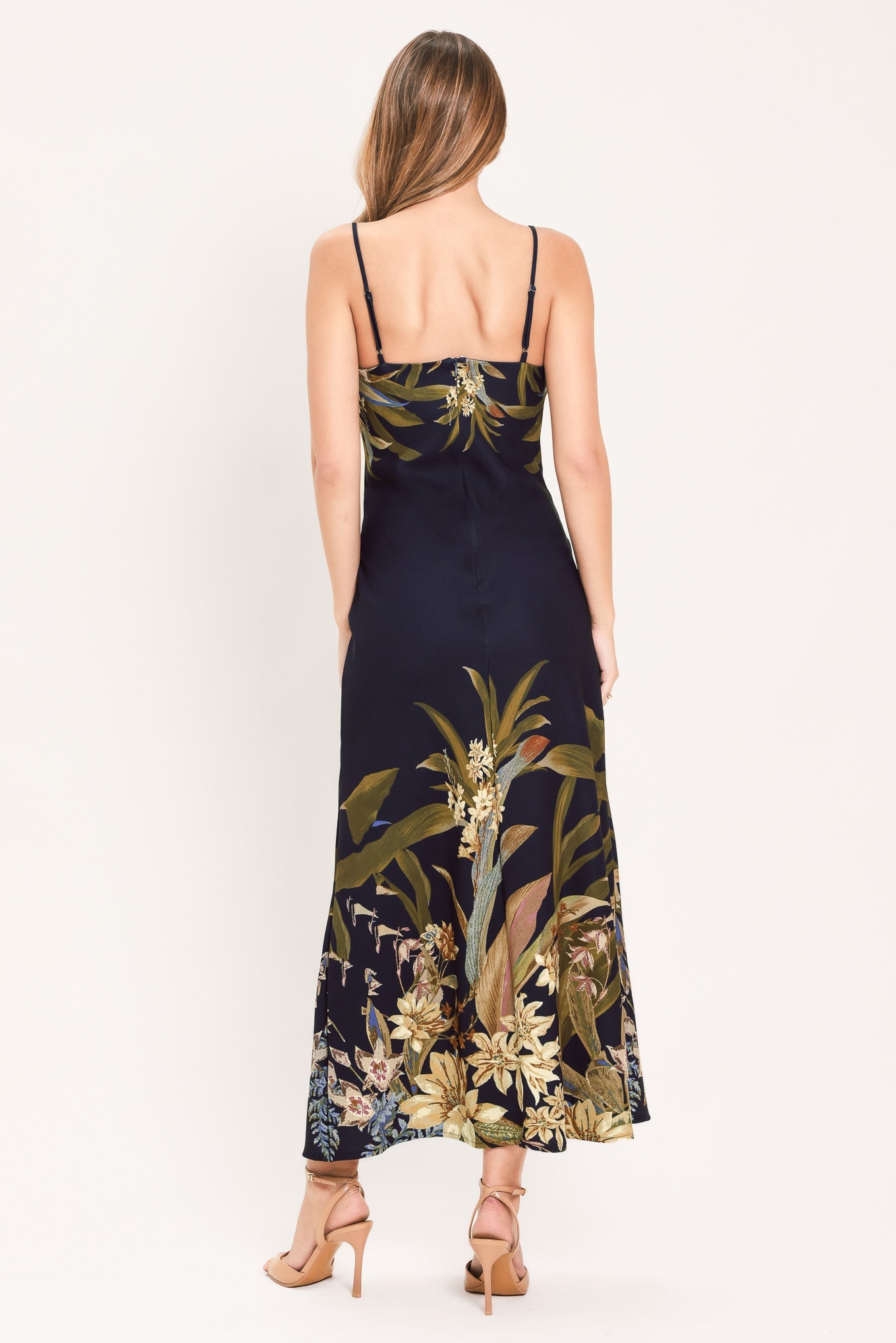 A woman shown from the back wearing a sleeveless maxi dress with thin straps and a dark navy base featuring a large botanical floral print in olive green, beige, and white. She is wearing beige high-heeled sandals against a plain white studio background.