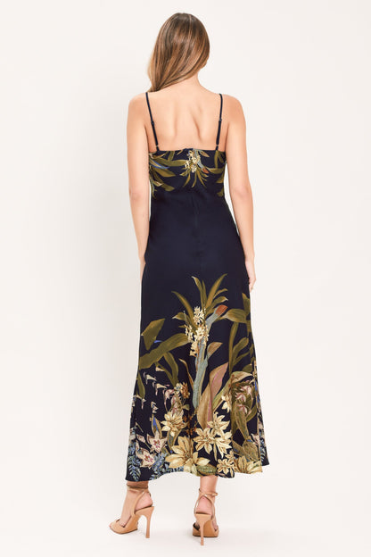 A woman shown from the back wearing a sleeveless maxi dress with thin straps and a dark navy base featuring a large botanical floral print in olive green, beige, and white. She is wearing beige high-heeled sandals against a plain white studio background.