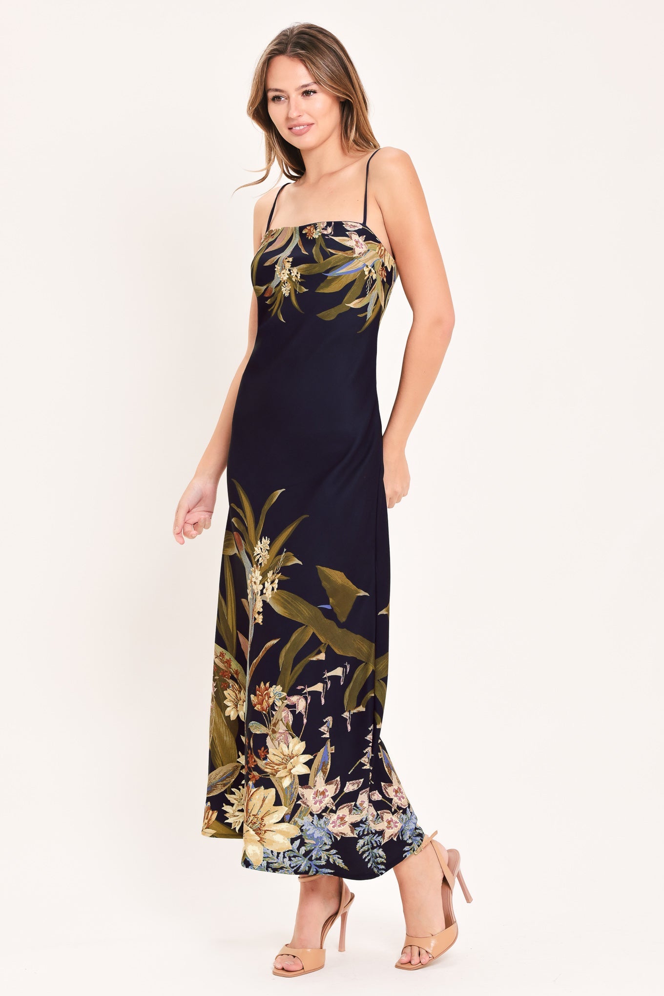 A woman modeling a navy maxi dress with a tropical floral and botanical print in olive green, beige, and light blue, paired with beige high-heeled sandals against a plain off-white studio background.