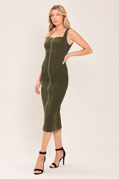 A woman modeling a fitted olive green midi dress with a square neckline, wide shoulder straps, and a central front zipper, paired with black high-heeled sandals against a plain light beige studio background.