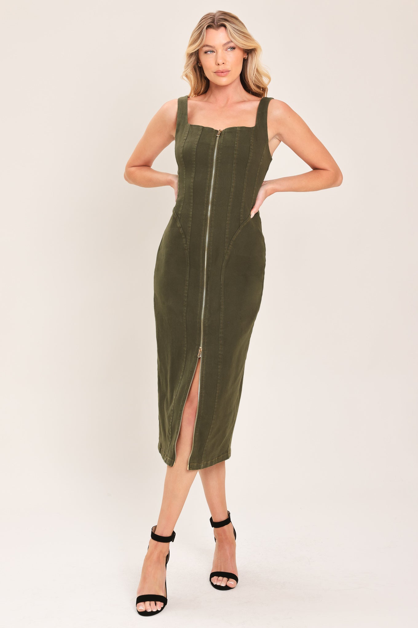 A woman modeling an olive green corduroy midi dress with a front zipper, square neckline, and side slit, paired with black high-heeled sandals against a plain light background.