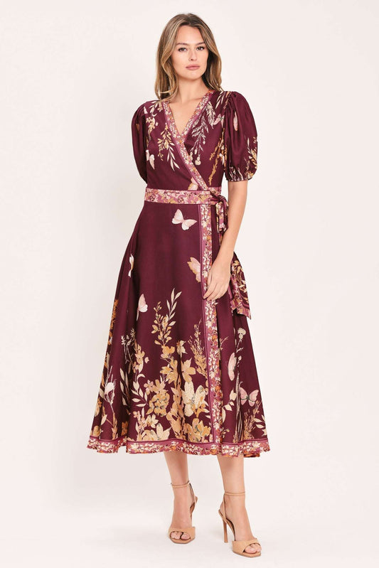 A woman modeling a midi wrap dress in deep burgundy with a gold and beige floral print, featuring puff sleeves and a matching waist tie, set against a plain light background.