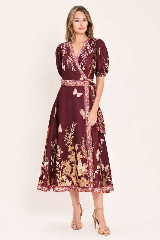 A woman modeling a midi wrap dress in deep burgundy with a gold and beige floral print, featuring puff sleeves and a matching waist tie, set against a plain light background.