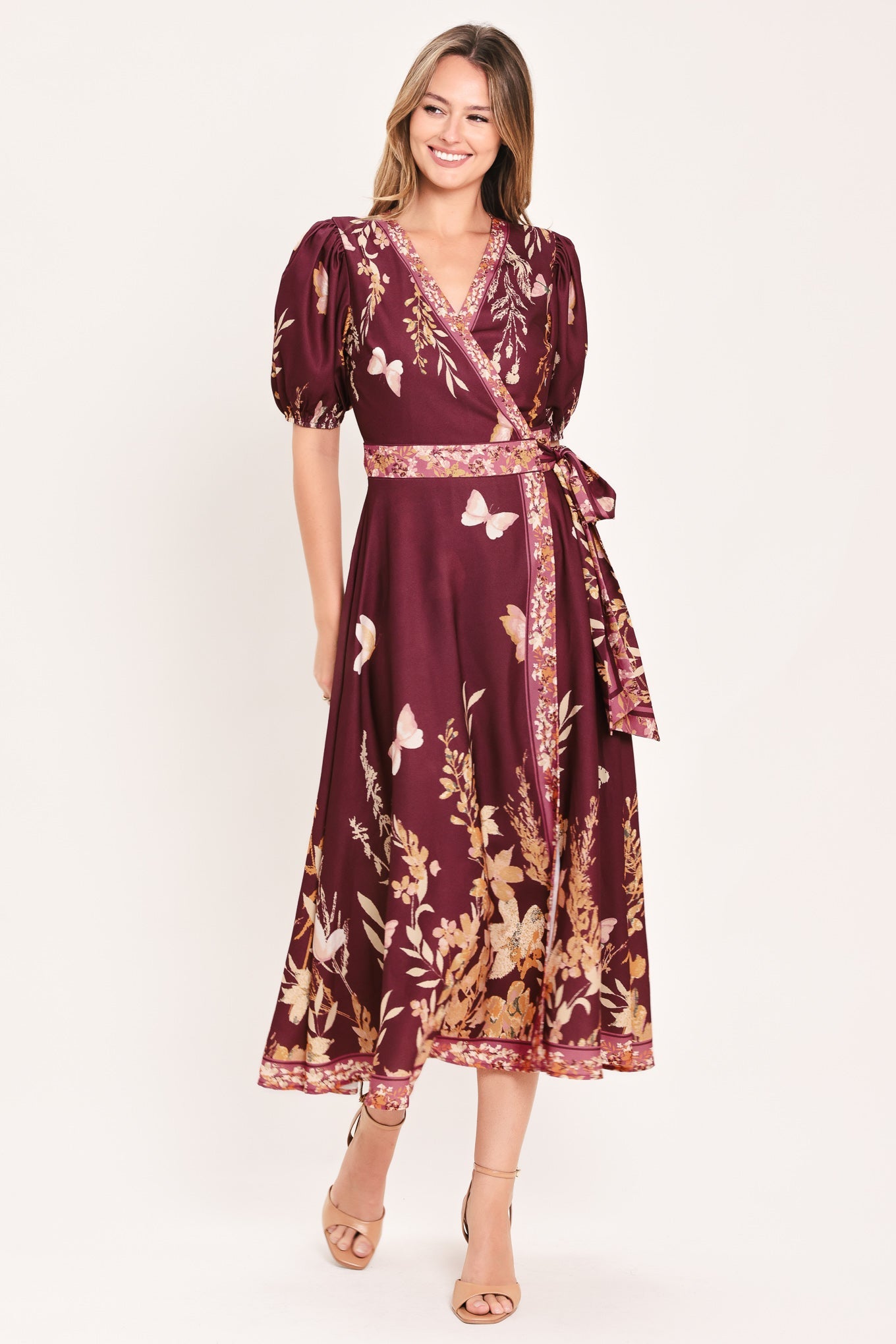 A woman modeling a burgundy midi wrap dress with a gold and white floral and butterfly print, featuring puff sleeves and a matching waist tie, set against a plain off-white studio background.