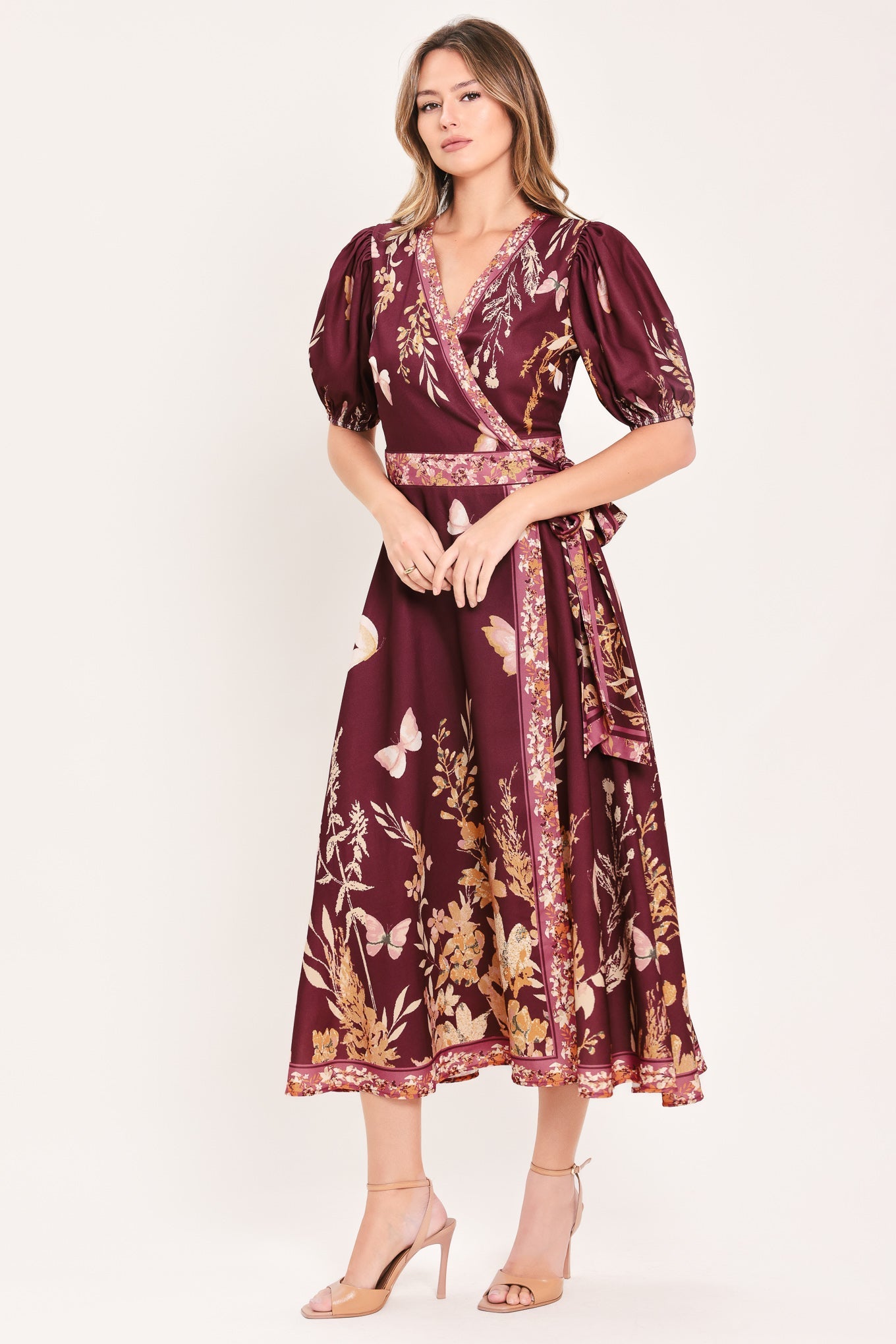 A woman modeling a burgundy midi wrap dress with a gold and beige floral and butterfly print, featuring puff sleeves and a matching waist tie, set against a plain light beige background.