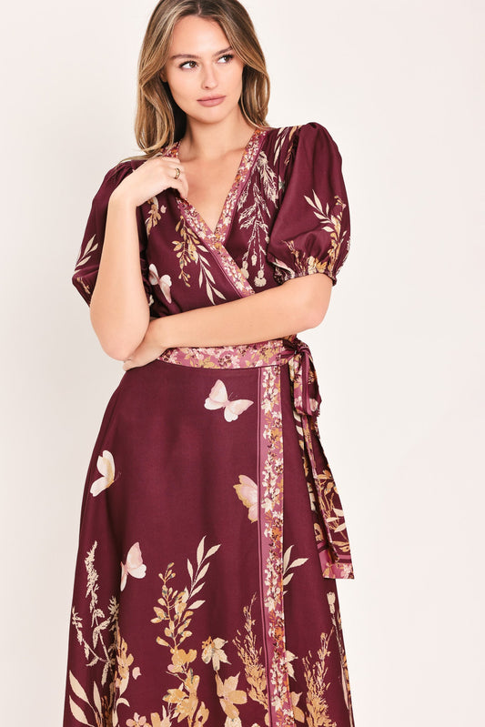 A woman wearing a deep burgundy wrap dress with a gold and white butterfly and floral print, set against a plain light beige background.