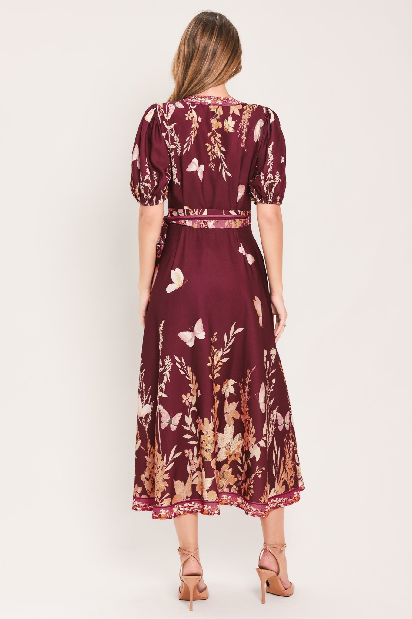 A woman viewed from the back wearing a burgundy midi dress with puff sleeves, a matching waistband, and a white and beige floral and butterfly print. She is wearing beige high-heeled sandals against a plain light background.