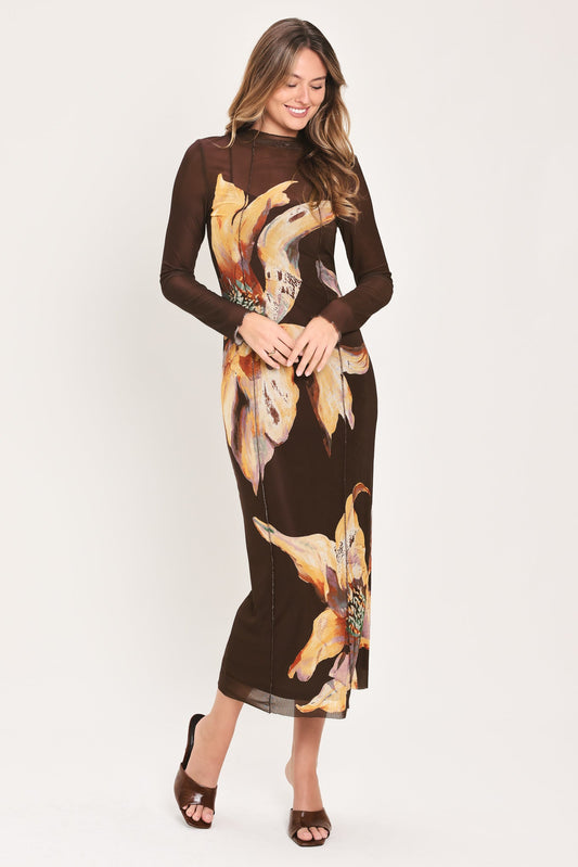 A woman modeling a long-sleeve brown midi dress with a large yellow and orange floral print, paired with brown heeled sandals, against a plain light background.