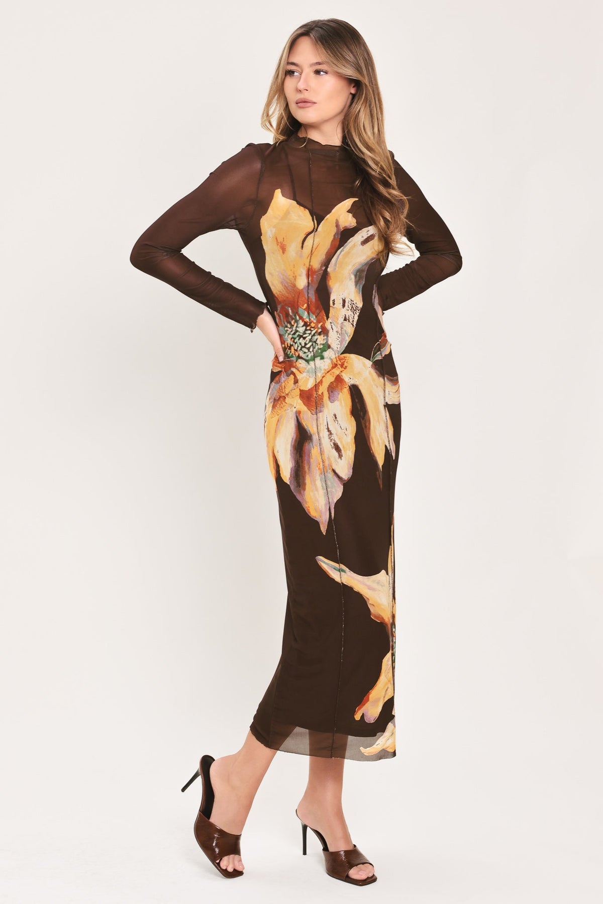 A woman modeling a long-sleeve brown midi dress with a sheer mesh overlay and a large yellow and orange floral print on the front, paired with brown heeled sandals against a light gray studio background.