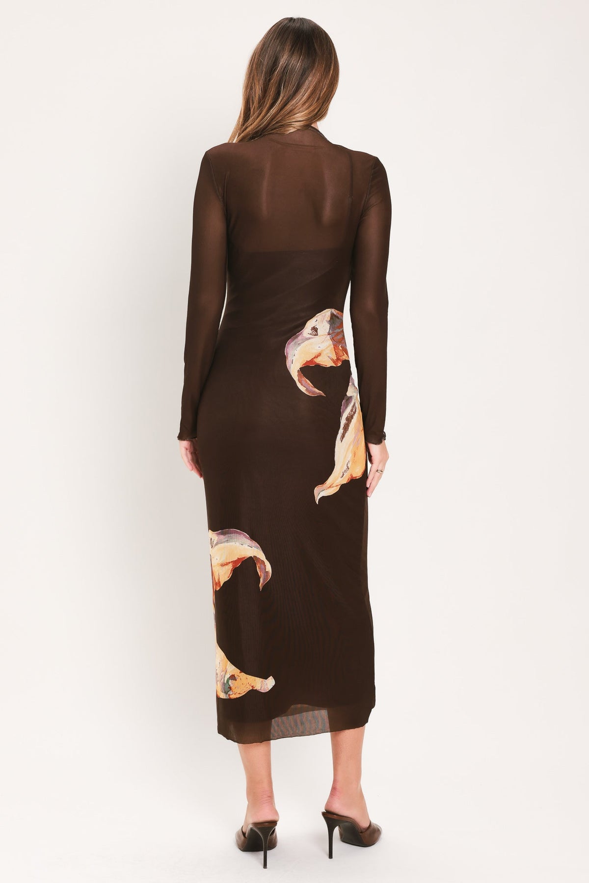 A woman viewed from the back wearing a dark brown long-sleeve midi dress with sheer mesh upper back and sleeves, plus scattered large tan and orange abstract leaf-like prints, paired with black heels against a plain light background.