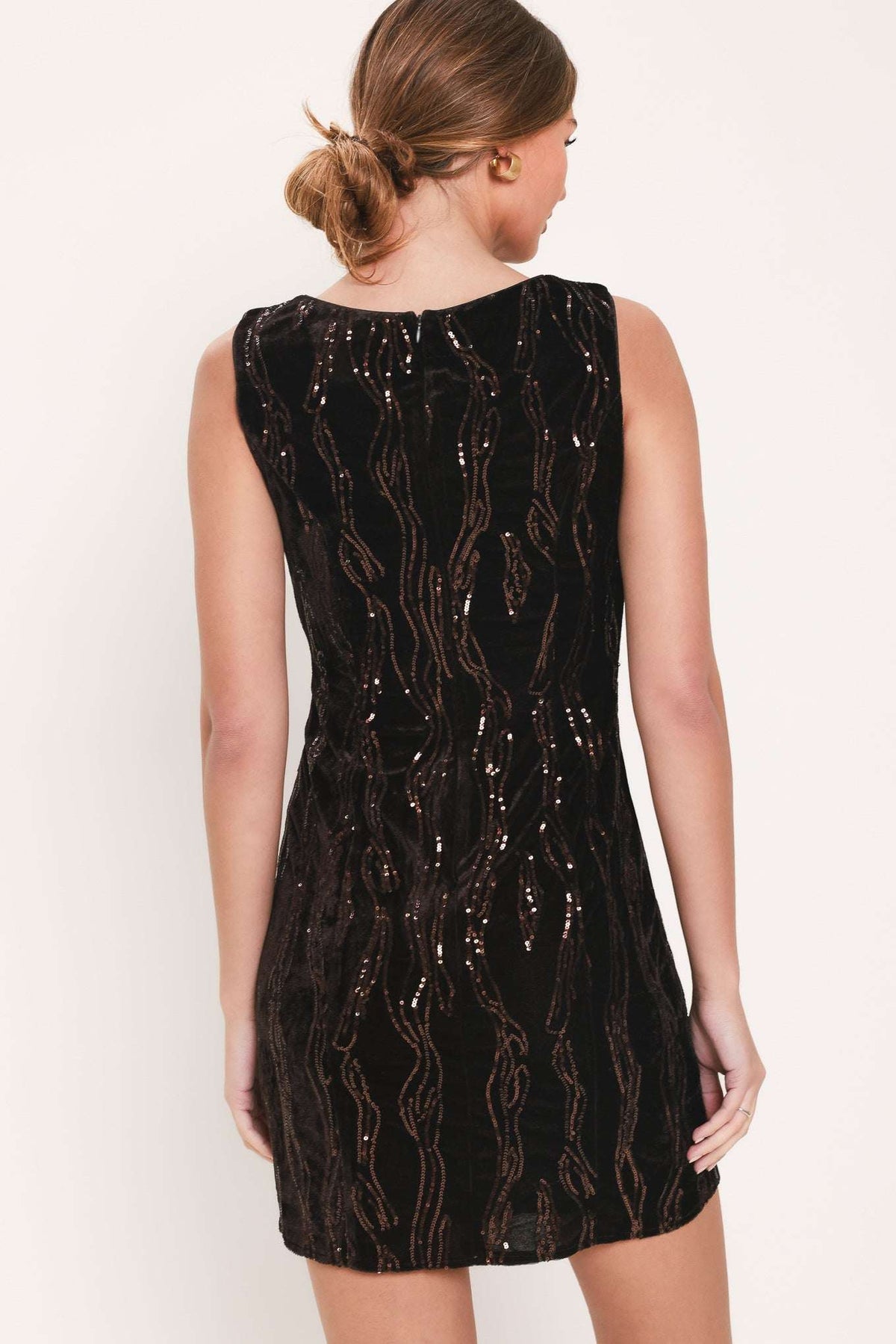 A woman shown from the back wearing a sleeveless black dress covered in vertical metallic gold sequin patterns against a plain off-white studio background.