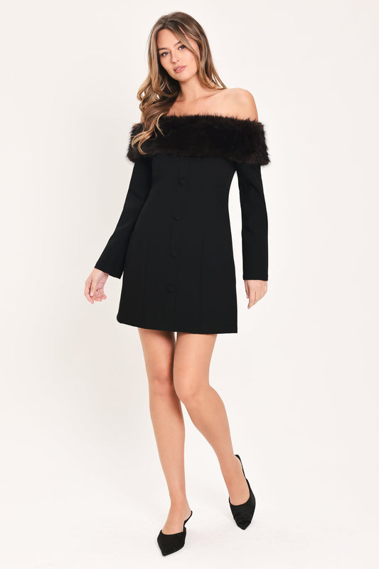 A woman modeling a black off-the-shoulder mini dress with a black faux-fur collar trim and front buttons, paired with black pointed-toe heels, against a plain light background.