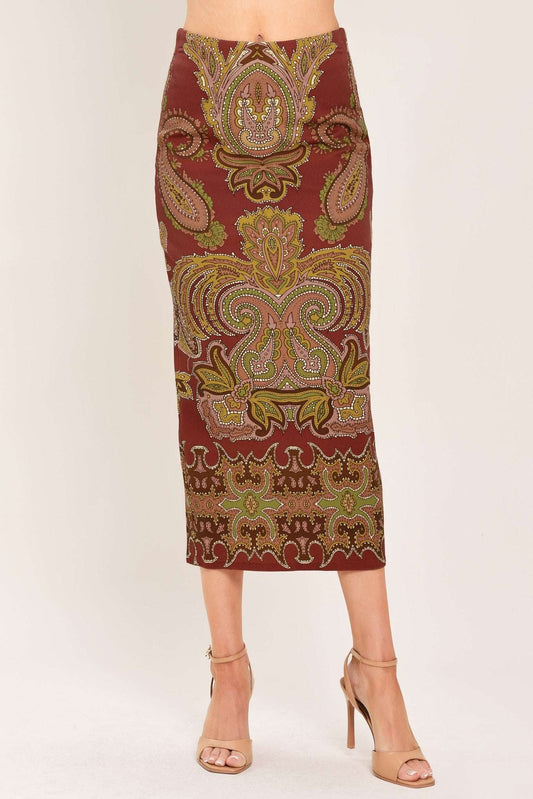 A woman models a knee-length fitted skirt with an ornate paisley pattern in rust red, gold, and olive green, paired with beige high-heeled sandals against a plain light background.