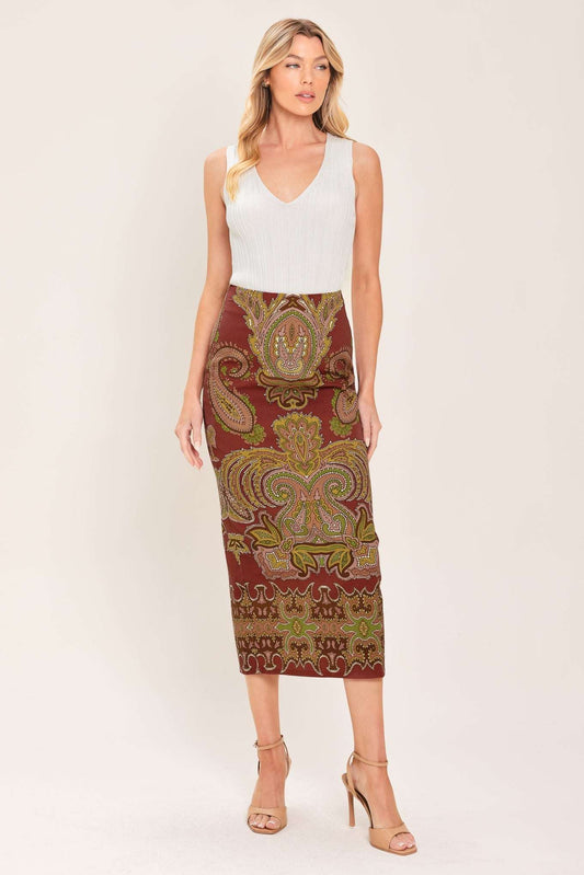 A woman modeling a sleeveless white V-neck top paired with a knee-length fitted skirt featuring an ornate paisley pattern in rust red, olive green, and gold, against a plain light beige studio background.