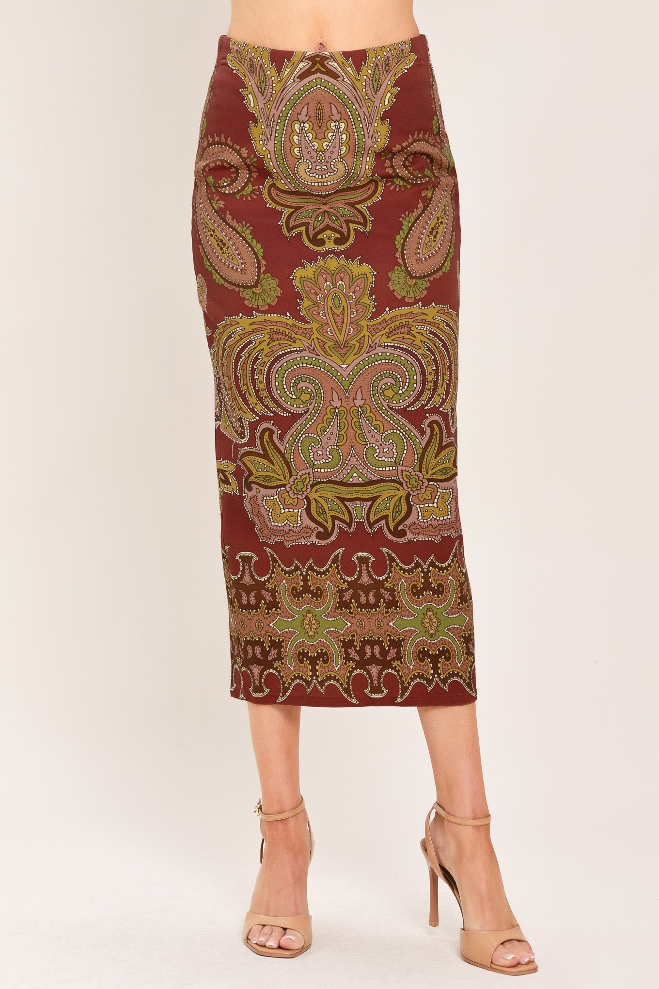 A woman models a knee-length fitted skirt with an ornate paisley pattern in rust red, gold, and olive green, paired with beige high-heeled sandals against a plain light background.
