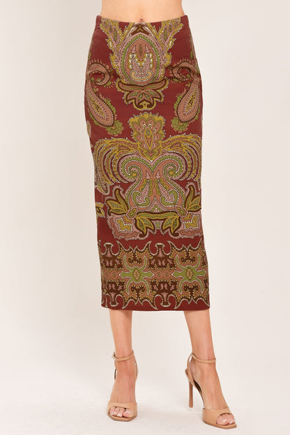 A woman models a knee-length fitted skirt with an ornate paisley pattern in rust red, gold, and olive green, paired with beige high-heeled sandals against a plain light background.