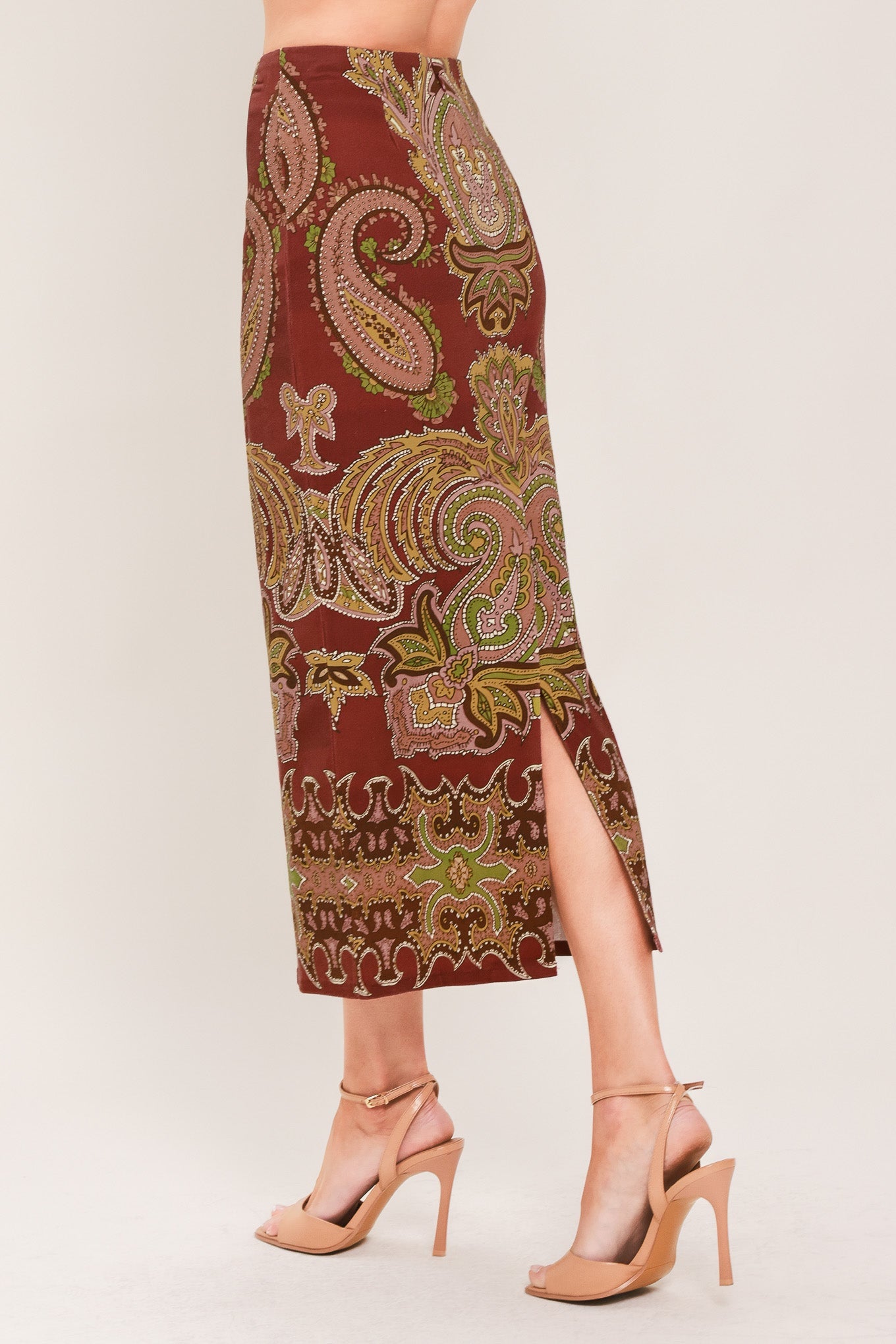A model's lower body wearing a rust-brown midi skirt with a green and gold paisley pattern, paired with nude high-heeled sandals, against a light beige studio background.