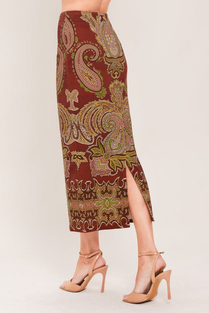 A model's lower body wearing a rust-brown midi skirt with a green and gold paisley pattern, paired with nude high-heeled sandals, against a light beige studio background.