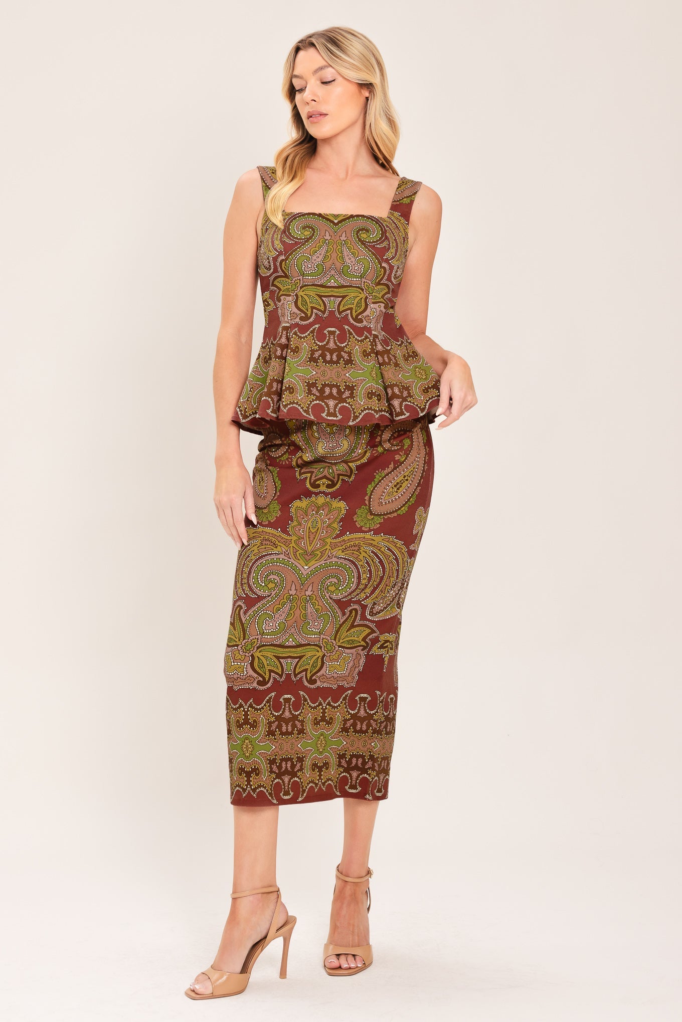 A woman modeling a sleeveless peplum midi dress with a brown, olive green, and gold paisley pattern, paired with nude high-heeled sandals, against a light beige studio background.