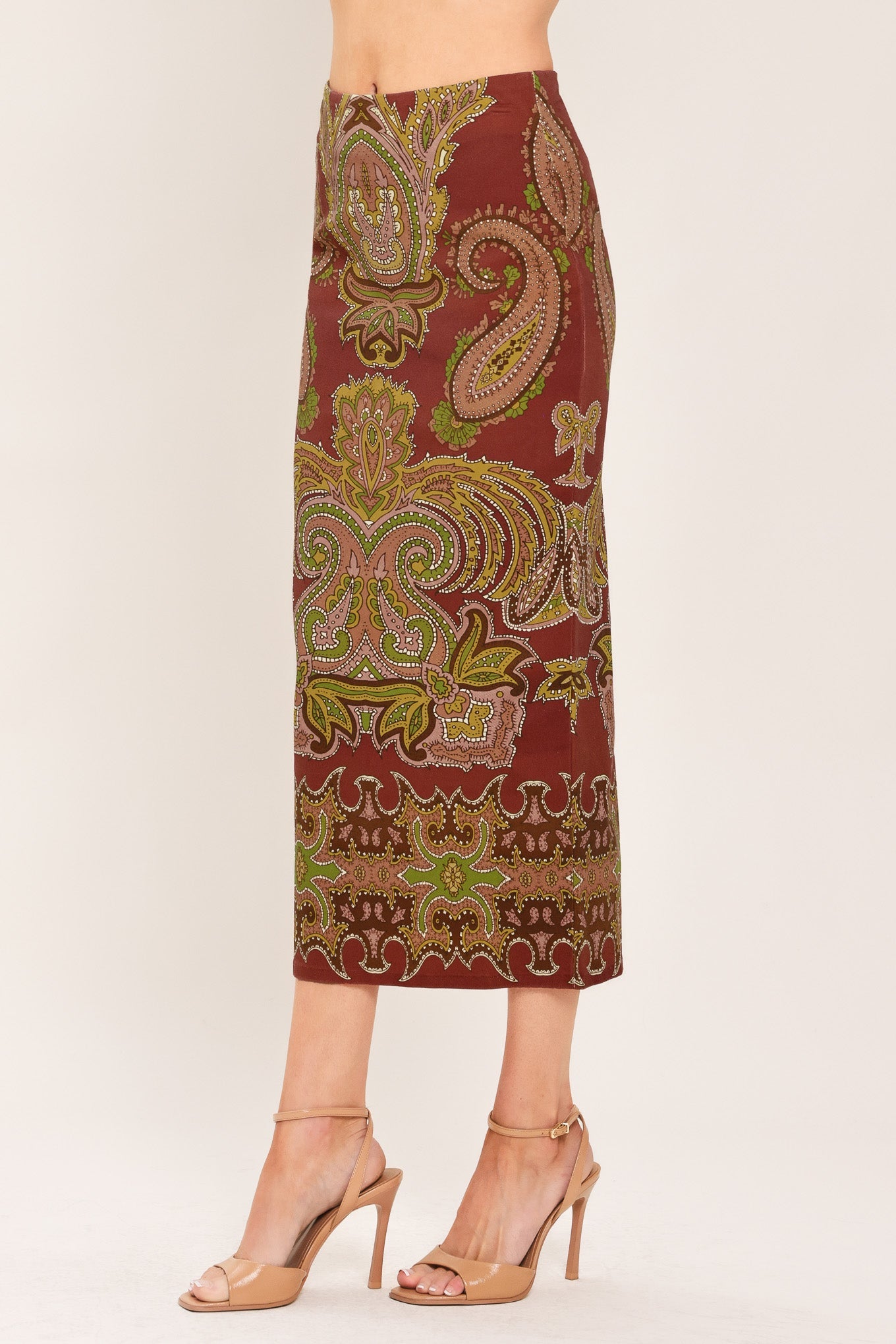 A model is wearing a mid-length skirt with an intricate paisley and floral pattern in rust red, olive green, and gold, paired with beige high-heeled sandals against a plain light background.
