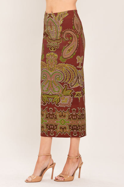 A model is wearing a mid-length skirt with an intricate paisley and floral pattern in rust red, olive green, and gold, paired with beige high-heeled sandals against a plain light background.