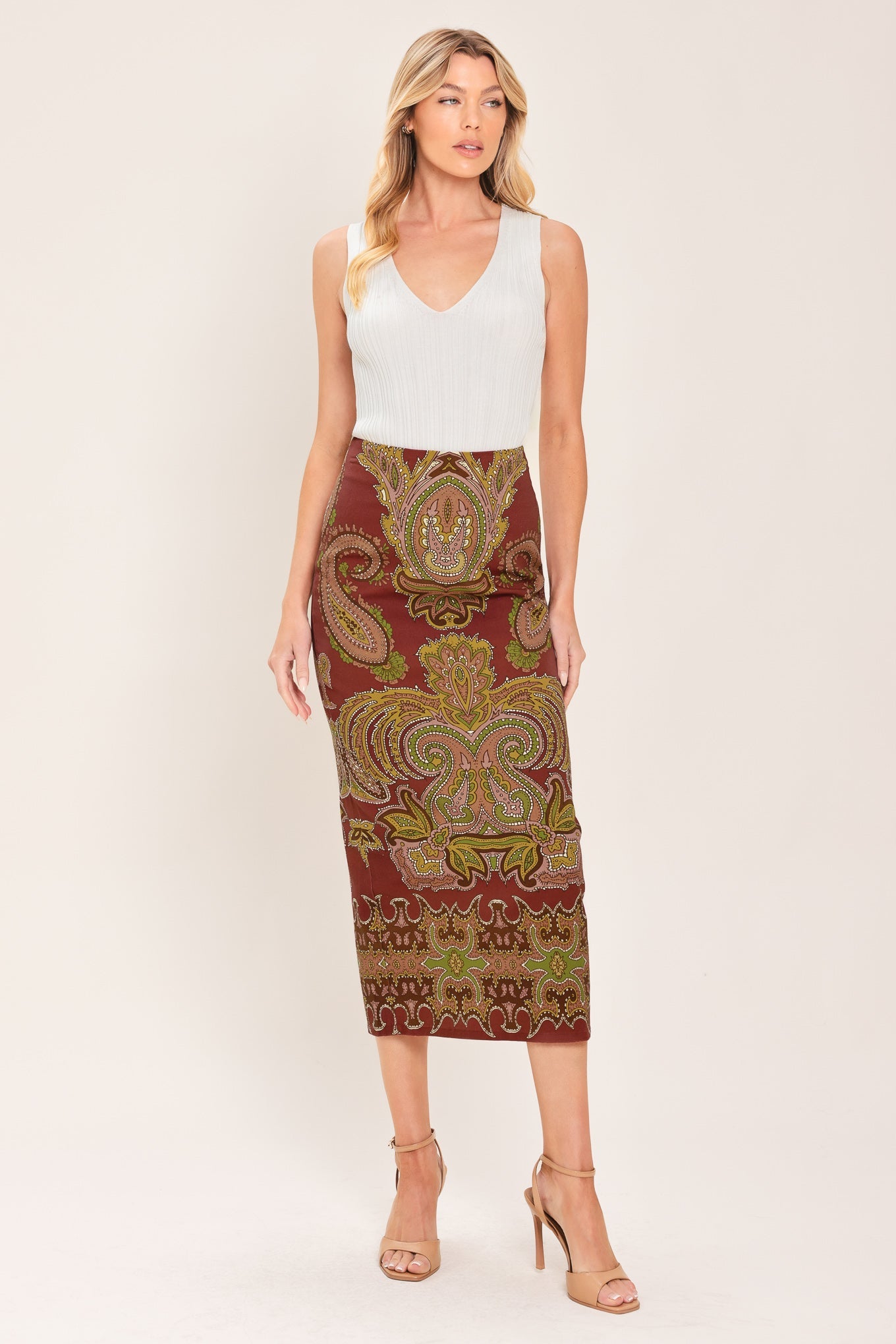 A woman modeling a sleeveless white V-neck top paired with a knee-length fitted skirt featuring an ornate paisley pattern in rust red, olive green, and gold, against a plain light beige studio background.