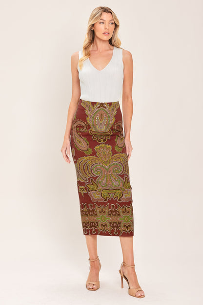 A woman modeling a sleeveless white V-neck top paired with a knee-length fitted skirt featuring an ornate paisley pattern in rust red, olive green, and gold, against a plain light beige studio background.