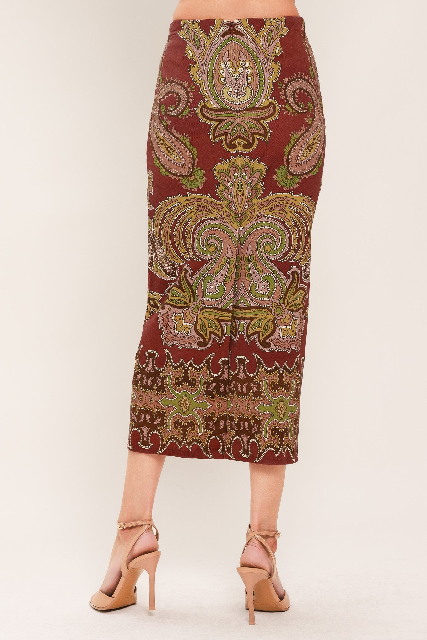 A woman wearing a knee-length fitted skirt with an ornate paisley and floral pattern in burgundy, gold, olive green, and cream, paired with tan high-heeled sandals against a plain light beige studio background.