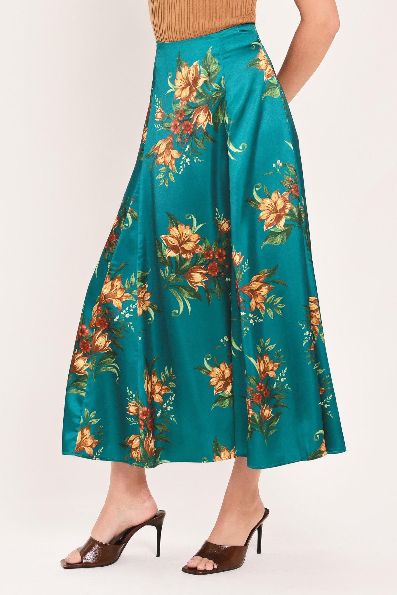 A model wearing a teal satin midi skirt with a vibrant orange and green floral print, paired with brown heeled sandals against a plain light background.