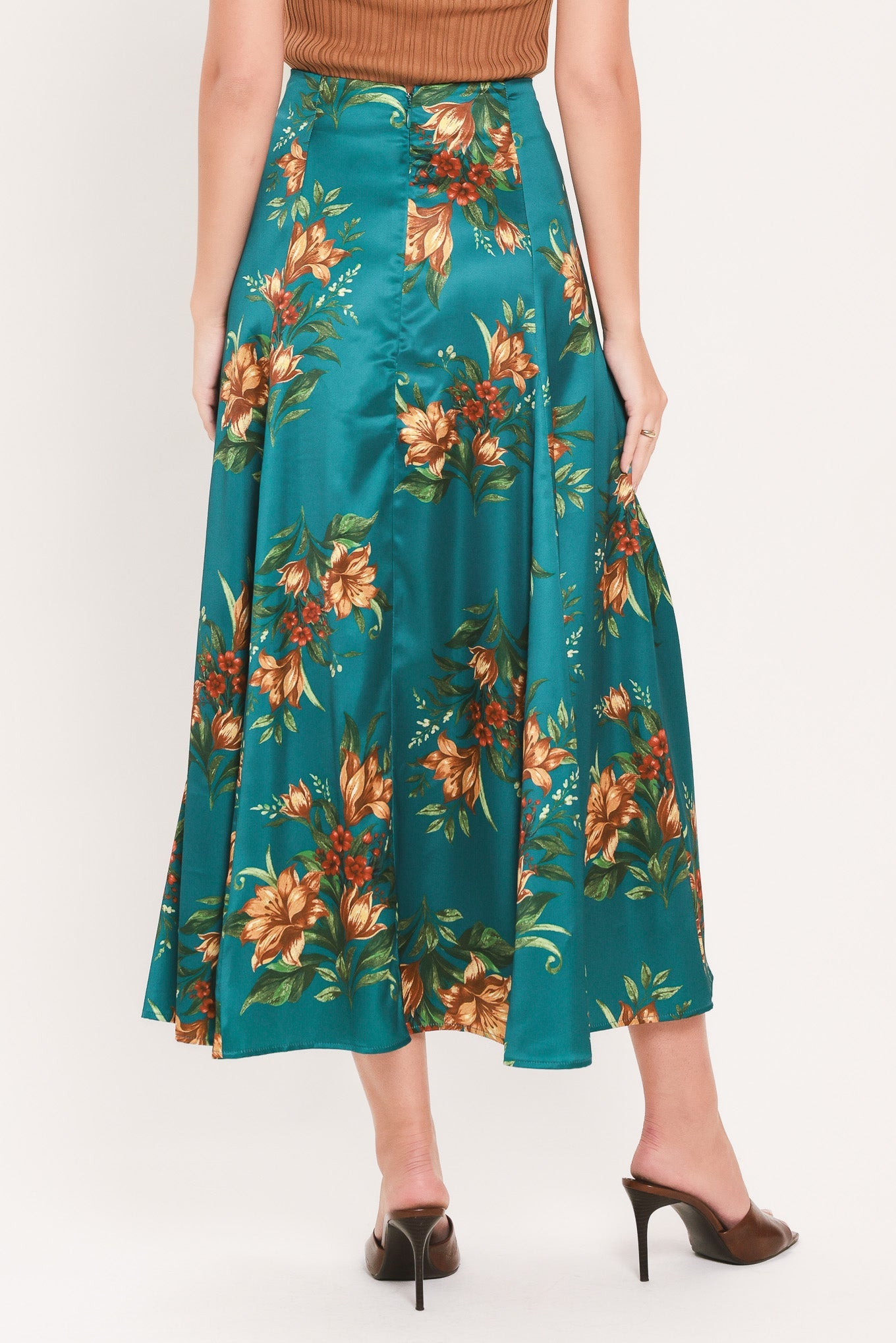 A model wearing a teal satin midi skirt with a large orange and green floral print, paired with a brown top and dark brown pointed-toe mule heels, shown from the waist down on a light gray studio background.
