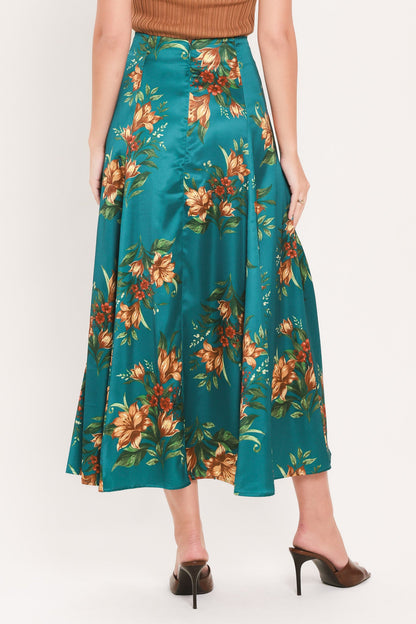 A model wearing a teal satin midi skirt with a large orange and green floral print, paired with a brown top and dark brown pointed-toe mule heels, shown from the waist down on a light gray studio background.