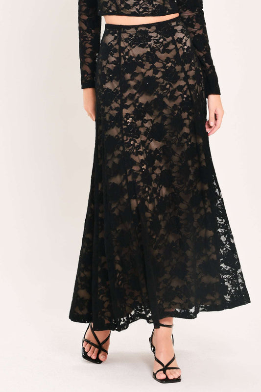 A model wearing a long black lace maxi skirt with a matching long-sleeve crop top, paired with black strappy high-heeled sandals, against a plain light background.
