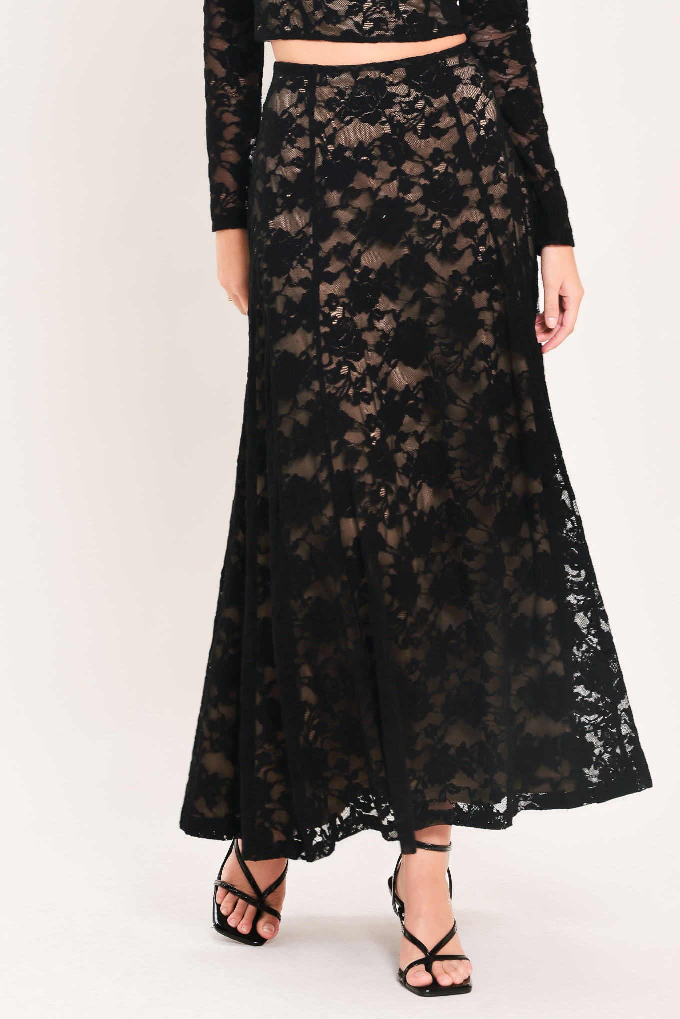 A model wearing a long black lace maxi skirt with a matching long-sleeve crop top, paired with black strappy high-heeled sandals, against a plain light background.