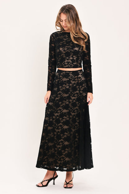 A woman modeling a black lace two-piece outfit consisting of a long-sleeve crop top and a matching maxi skirt, paired with black strappy heels, against a plain white studio background.