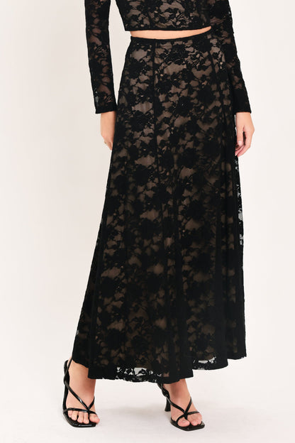 A model wearing a black lace two-piece set consisting of a long-sleeve crop top and a matching high-waisted maxi skirt, paired with black strappy heels, against a plain light background.
