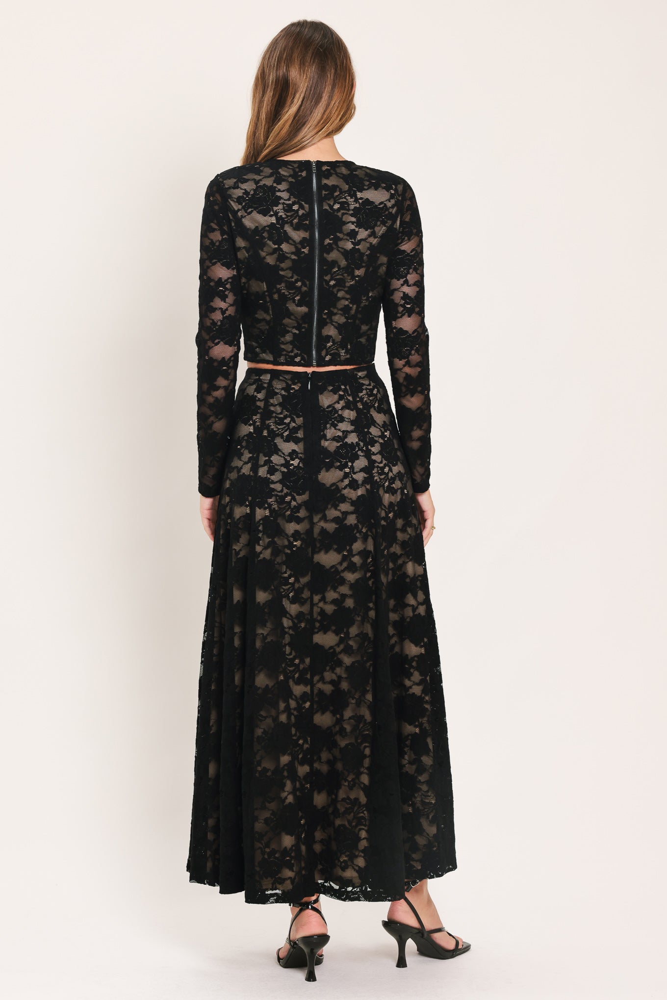 A woman viewed from the back wearing a long-sleeve black lace maxi dress with a thin brown belt at the waist, paired with black heels, against a plain light beige studio background.
