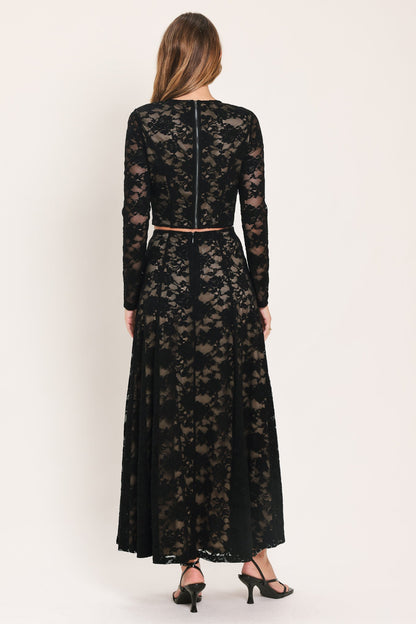 A woman viewed from the back wearing a long-sleeve black lace maxi dress with a thin brown belt at the waist, paired with black heels, against a plain light beige studio background.