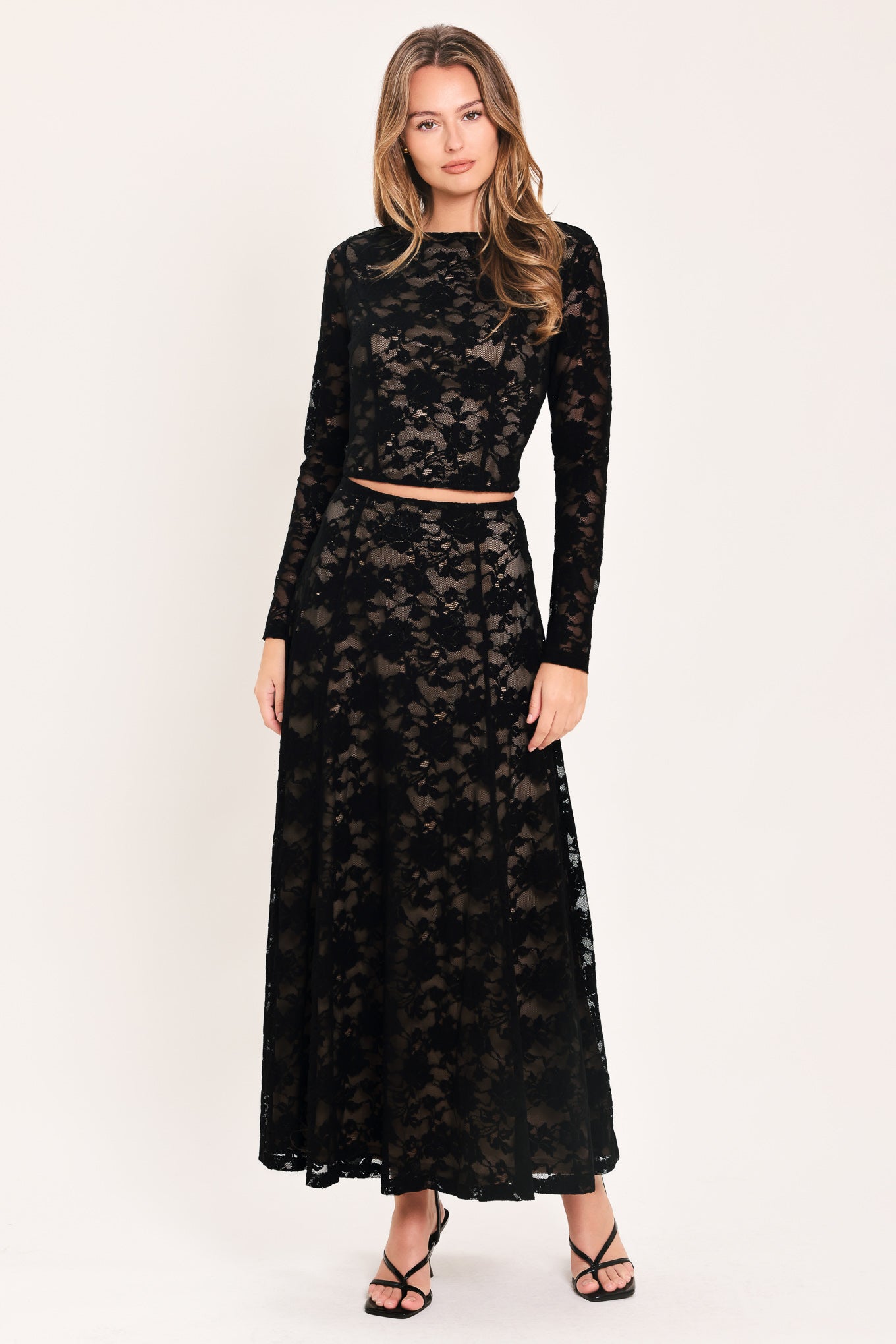 A woman modeling a black lace two-piece outfit consisting of a long-sleeve crop top and a matching high-waisted maxi skirt, paired with black strappy heels against a plain off-white studio background.