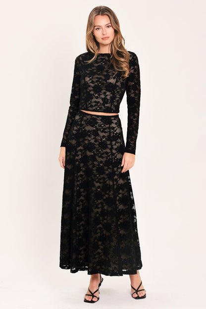A woman modeling a black lace two-piece outfit consisting of a long-sleeve crop top and a matching high-waisted maxi skirt, paired with black strappy heels against a plain off-white studio background.