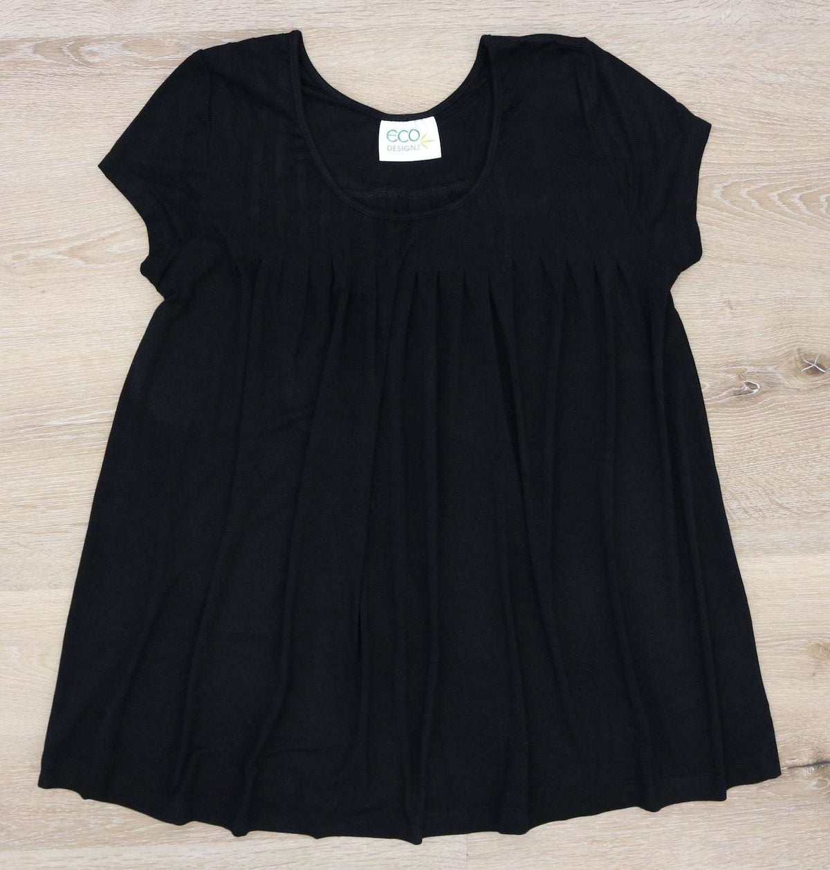 Black short-sleeve pleated fabric top with an ECO DESIGNZ label, laid flat on a light wood surface.