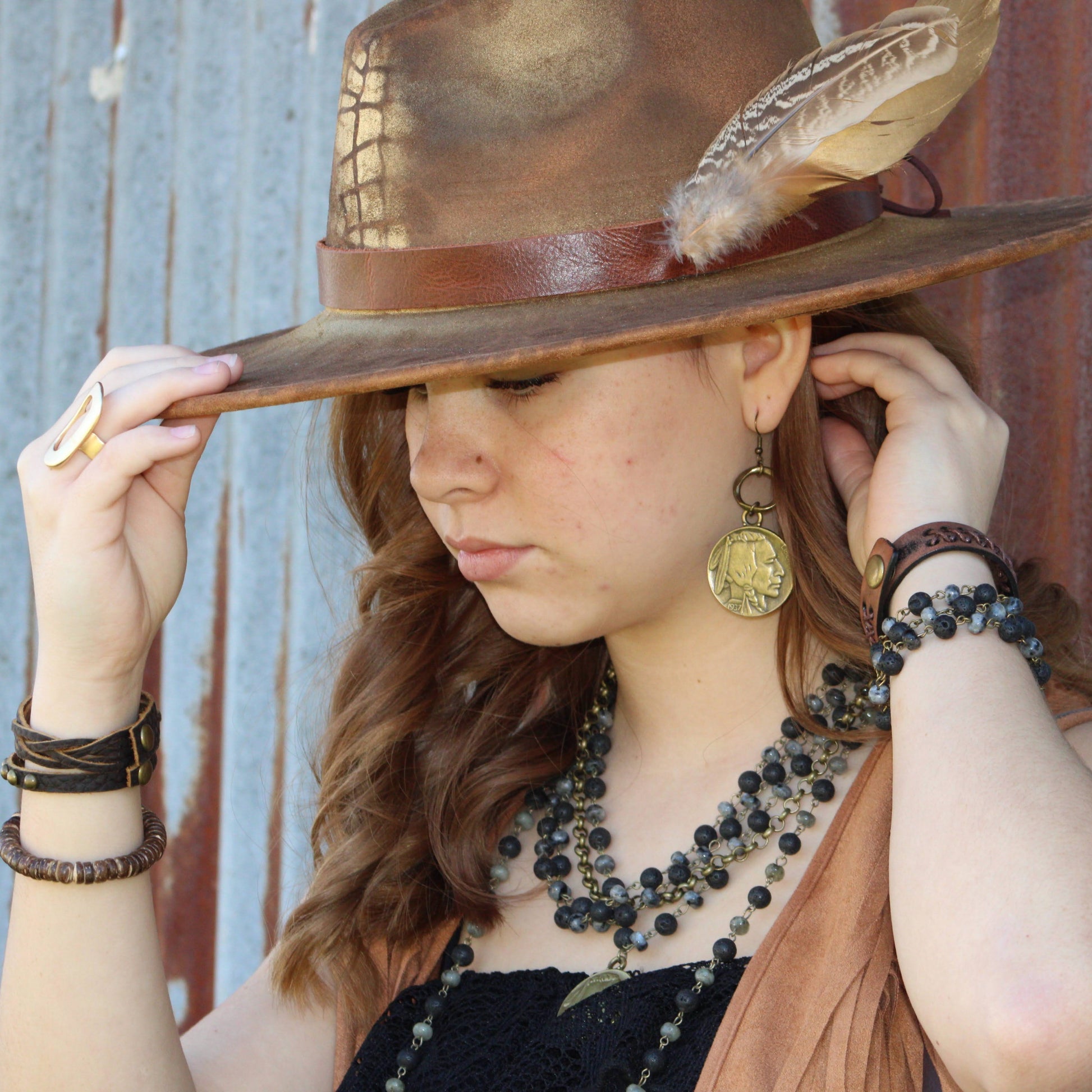 Woman wearing a wide-brimmed hat with a feather, multiple necklaces, and bracelets against a rustic wooden background.