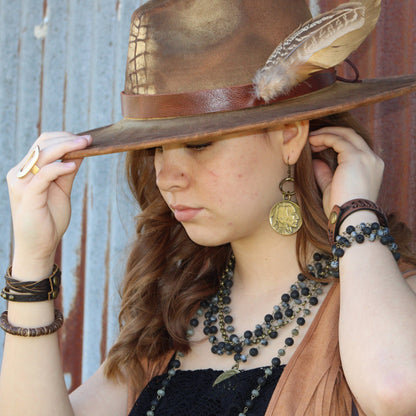 Woman wearing a wide-brimmed hat with a feather, multiple necklaces, and bracelets against a rustic wooden background.