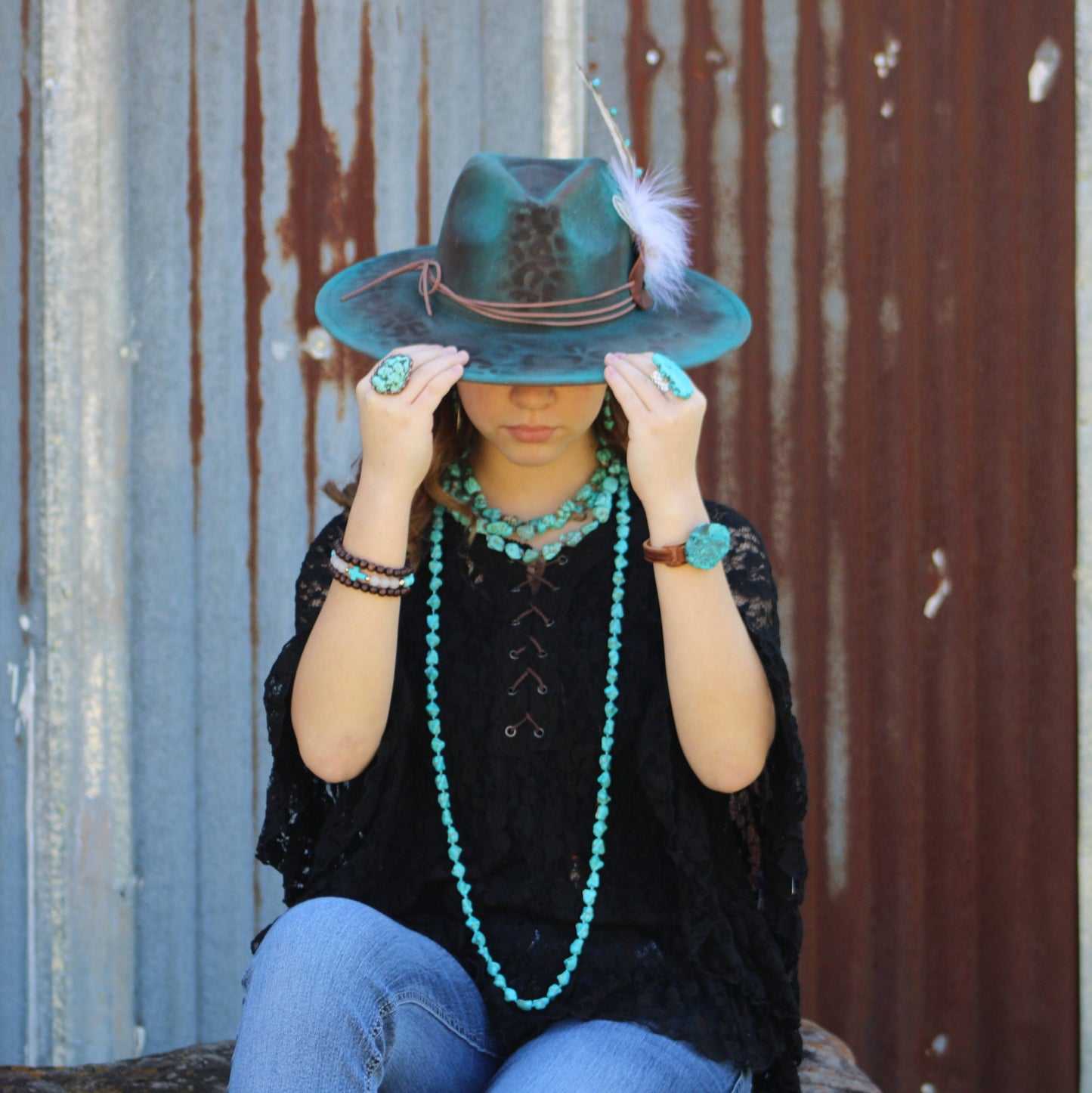 Person wearing a turquoise hat with a feather, black top, and blue jeans, sitting against a rusty metal wall.